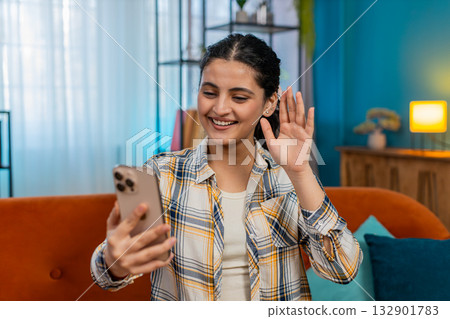 Indian woman on sofa happily waving hand greeting via video call on smartphone smiling joyful 132901783
