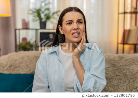 Young woman sitting on sofa holding cheek suffering from strong toothache feeling pain uncomfortable 132901786