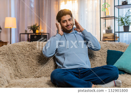 Indian young man sitting on sofa listening to music with headphones dancing to rhythm at home 132901800