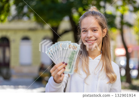 Happy Caucasian young woman girl holding fan of dollars showing thumbs up gesture on city street 132901914