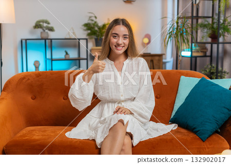 Portrait of excited smiling young woman showing double thumbs up sitting on sofa couch at home 132902017