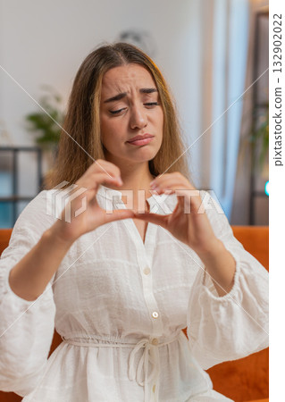 Portrait of upset young woman in white dress sitting on sofa showing heartbroken sign with her hands 132902022