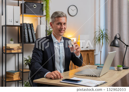Handsome young freelancer using laptop and drinking glass of water sitting at home office desk Handsome young freelancer using laptop and drinking glass of water sitting at home office desk 132902033