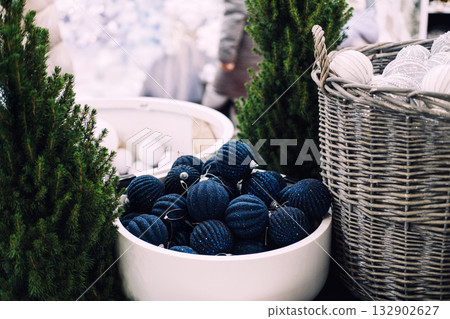 Blue glittery Christmas ornaments in a white bowl displayed between artificial pine trees and wicker baskets. Bulk holiday decor, snowball ornaments wholesale, retail-ready seasonal props Blue glittery Christmas ornaments in a white bowl displayed between artificial pine trees and wicker baskets. Bulk holiday decor, snowball ornaments wholesale, retail-ready seasonal props 132902627