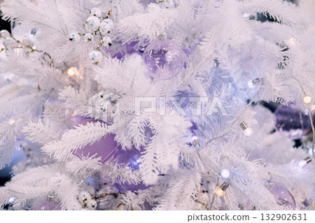Close-up of a white artificial Christmas tree decorated with pastel purple ornaments, faux berries, and string lights. Pastel holiday styling, dreamy winter decor, artificial tree closeup 132902631