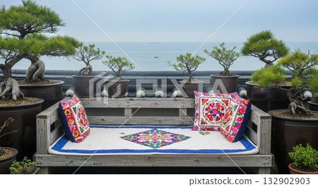 Serene oceanfront terrace with decorative bonsai pine trees in pottery bowls and colorful embroidered cushions on weathered wooden bench overlooking calm blue sea 132902903