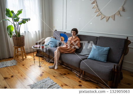 Woman suffering from overheating at home, waving hand paper fan and scrolling media on smartphone. 132905063