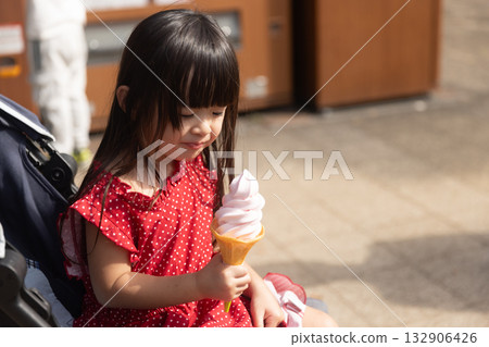 一個穿著紅色洋裝的3歲女孩坐在嬰兒車裡吃軟冰淇淋 132906426