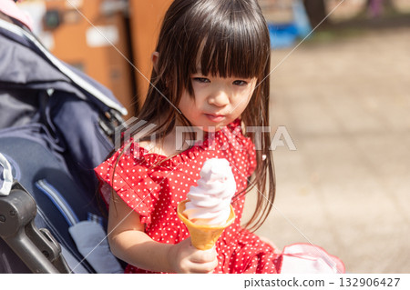 A 3-year-old girl in a red dress sitting in a stroller and eating soft serve ice cream 132906427