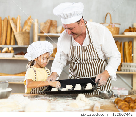 Little girl in striped apron and white chefs hat near kitchen table puts croissants on baking sheet Little girl in striped apron and white chefs hat near kitchen table puts croissants on baking sheet 132907442