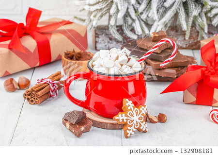Red mug of hot chocolate with marshmallows and candy cane surrounded by gingerbread cookie, chocolate pieces, cinnamon sticks and Christmas gifts on festive white background Red mug of hot chocolate with marshmallows and candy cane surrounded by gingerbread cookie, chocolate pieces, cinnamon sticks and Christmas gifts on festive white background 132908181
