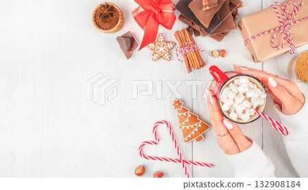female hands holding red mug of hot chocolate with marshmallows and candy cane surrounded by gingerbread cookies, chocolate, cinnamon, and festive gifts on light background copy space. Top view female hands holding red mug of hot chocolate with marshmallows and candy cane surrounded by gingerbread cookies, chocolate, cinnamon, and festive gifts on light background copy space. Top view 132908184
