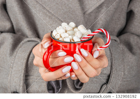 Woman in gray coat holding red mug of hot chocolate with marshmallows and candy cane, cozy winter holiday mood close-up Woman in gray coat holding red mug of hot chocolate with marshmallows and candy cane, cozy winter holiday mood close-up 132908192