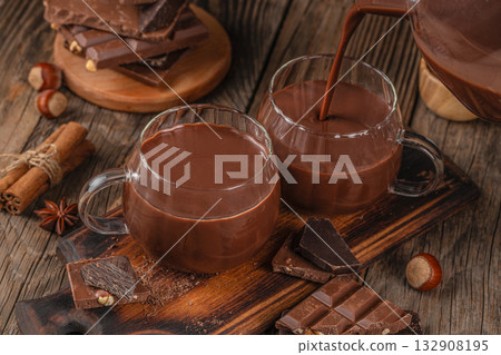 Hot chocolate being poured into glass mugs with pieces of dark and milk chocolate, cinnamon sticks, and hazelnuts on rustic wooden background, cozy winter beverage concept Hot chocolate being poured into glass mugs with pieces of dark and milk chocolate, cinnamon sticks, and hazelnuts on rustic wooden background, cozy winter beverage concept 132908195