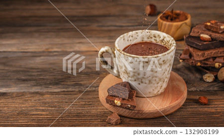 Cup of hot chocolate on wooden table with falling cocoa powder, pieces of dark and milk chocolate with hazelnuts, cozy rustic background with copy space for warm drink concept 132908199