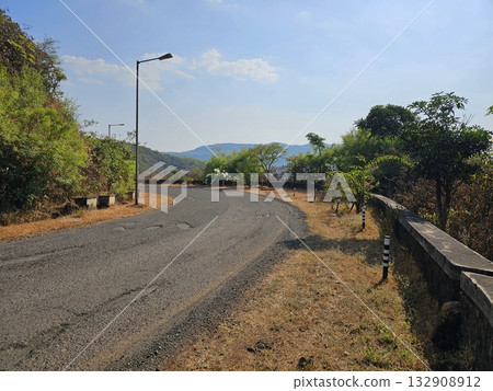 Curving hill road Empty winding road bordered by trees and streetlights on a sunny hillside, symbolizing freedom, solitude, and exploration. 132908912