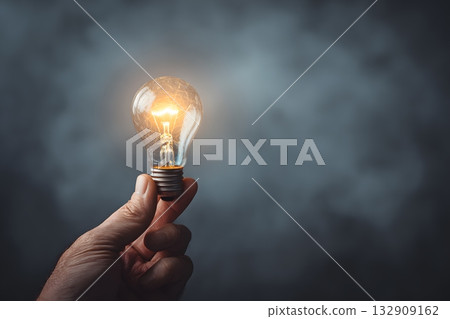 A glowing light bulb is held in a hand against a dark, smoky background. 132909162