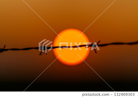 A Barbed Wire Fence Silhouetted Against a Beautiful and Vibrant Sunset in the Sky 132909303