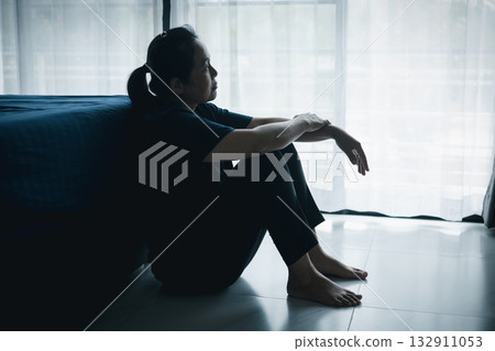 A lonely woman sits on the floor by window looking outside in dark room. Concept of depression, anxiety, sadness, loneliness, mental health problem, emotional struggle and hope for better life. A lonely woman sits on the floor by window looking outside in dark room. Concept of depression, anxiety, sadness, loneliness, mental health problem, emotional struggle and hope for better life. 132911053