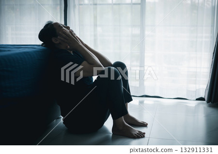 A lonely woman sits on the floor by window with hand on head in dark room. Concept of depression, sadness, anxiety, stress, mental health problem, loneliness and emotional struggle in life. 132911331