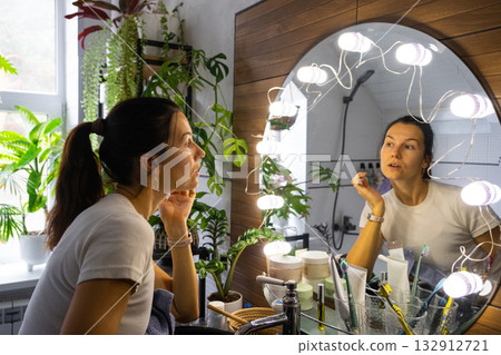 A woman washes her face in the bathroom and looks in the illuminated mirror. Morning and evening hygiene A woman washes her face in the bathroom and looks in the illuminated mirror. Morning and evening hygiene 132912721