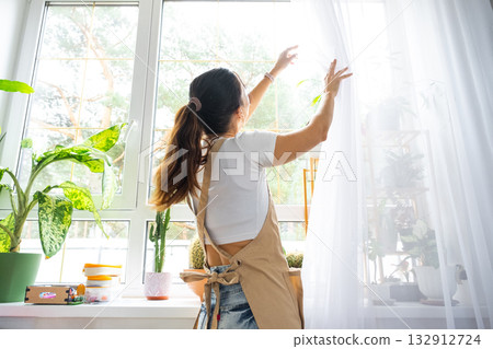 Woman in apron hangs transparent tulle curtains on large windows in the house inside the interior with potted plants. Spring cleaning, tidying up 132912724
