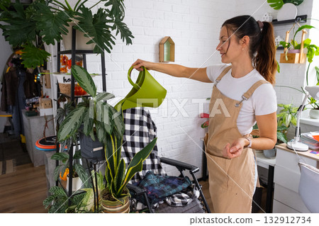 A woman waters home plants from her collection of rare species from a watering can, grown with love on shelves in the interior of the house. Home plant growing, green house, water balance A woman waters home plants from her collection of rare species from a watering can, grown with love on shelves in the interior of the house. Home plant growing, green house, water balance 132912734