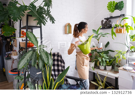 A woman waters home plants from her collection of rare species from a watering can, grown with love on shelves in the interior of the house. Home plant growing, green house, water balance A woman waters home plants from her collection of rare species from a watering can, grown with love on shelves in the interior of the house. Home plant growing, green house, water balance 132912736