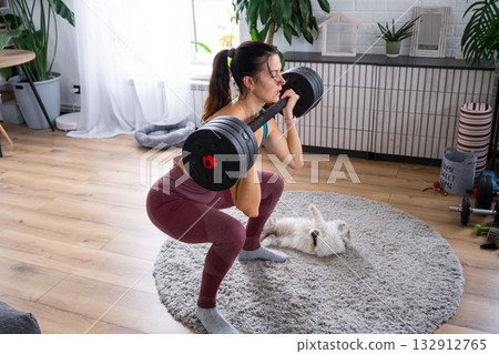 A woman in a sports uniform is engaged in fitness and strength training with a barbell and dumbbells at home in the interior and cat A woman in a sports uniform is engaged in fitness and strength training with a barbell and dumbbells at home in the interior and cat 132912765