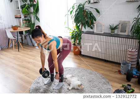 A woman in a sports uniform is engaged in fitness and strength training with a barbell and dumbbells at home in the interior and cat 132912772