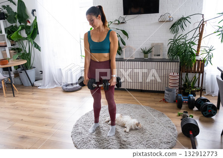 A woman in a sports uniform is engaged in fitness and strength training with a barbell and dumbbells at home in the interior and cat 132912773