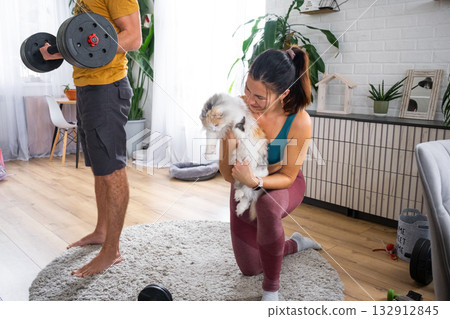 Woman and man in a sports uniform is engaged in fitness and strength training with a barbell and dumbbells at home in the interior and cat 132912845
