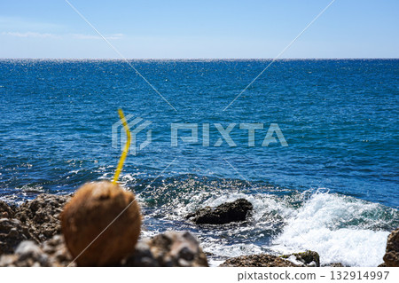 A Refreshing Coconut Drink Set Against a Stunning Ocean View with Waves Crashing on Rocky Shore Under a Clear Blue Sky 132914997