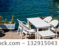 A Tranquil Waterfront Scene Featuring White Furniture and Calm Waters Surrounded by Lush Greenery and Colorful Flowers 132915043