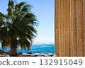 A Scenic View of Ancient Columns and Lush Palm Trees Overlooking a Tranquil Ocean Under a Bright Blue Sky 132915049