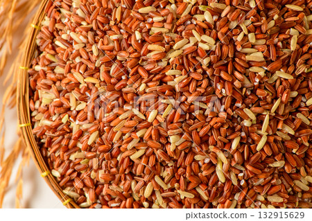 Close up of organic brown rice grains in a bamboo basket with golden rice stalks on a white background. 132915629
