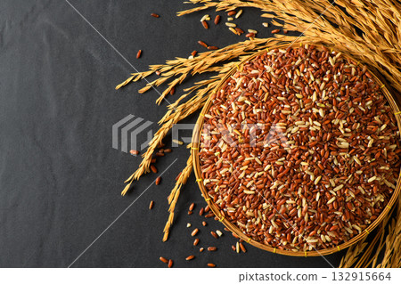 Close up of organic brown rice grains in a bamboo basket with golden rice stalks on black stone 132915664