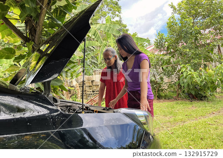 An elderly and young Asian woman looks under the hood of a broken down car to find a problem with the engine An elderly and young Asian woman looks under the hood of a broken down car to find a problem with the engine 132915729