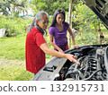 An elderly and young Asian woman looks under the hood of a broken down car to find a problem with the engine 132915731