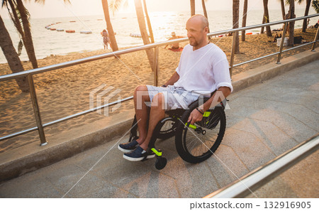 Disabled man in a wheelchair moves on a ramp to the beach. Disabled man in a wheelchair moves on a ramp to the beach. 132916905