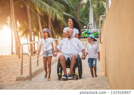 Disabled man in a wheelchair moves on a ramp to the beach with his family. 132916907