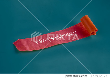 A vibrant piece of textured paper rolls out across a solid blue background, showcasing the bold phrase always hungry in playful handwriting. The warm colors invite curiosity and creativity 132917125