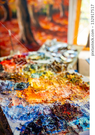 Close-up of a messy painter palette with thick dabs of oil paint in various bright colors. In the background, a blurry painting is visible, evoking creativity and inspiration. Close-up of a messy painter palette with thick dabs of oil paint in various bright colors. In the background, a blurry painting is visible, evoking creativity and inspiration. 132917517
