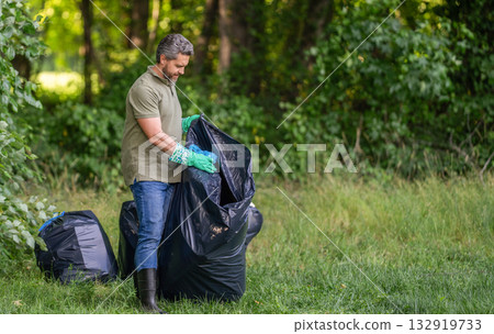 Man in rubber gloves with trash bag clean up garbage. Man collecting plastic. Eco volunteer. Garbage collecting in forest. Ecology concept. Volunteer collecting garbage, ecology. Social advertisement 132919733