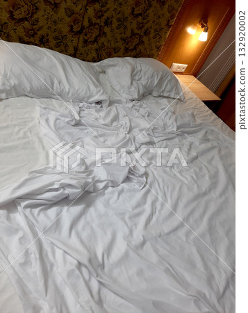 Crumpled white bed sheets on a neatly made bed with warm lighting and floral wallpaper Crumpled white bed sheets on a neatly made bed with warm lighting and floral wallpaper 132920002