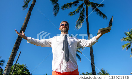 Businessman successful man using laptop under palm tree at business trip having success in freelance work online in summer vacation Businessman successful man using laptop under palm tree at business trip having success in freelance work online in summer vacation 132921586