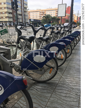 Row of bicycles at public rental station on city street in Valencia. Mobility, sustainability, and modern urban transportation for eco-friendly lifestyle and daily movement. Row of bicycles at public rental station on city street in Valencia. Mobility, sustainability, and modern urban transportation for eco-friendly lifestyle and daily movement. 132921677