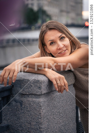Woman in a knitted dress leans on a stone railing by the river at dusk, looking into the camera with a soft smile 132921866