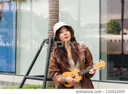 Young woman playing ukulele, happy person enjoying summer travel holiday, feeling joyful, sharing music on outdoor city street, stylish young tourist with casual fashion, long brown hair. Young woman playing ukulele, happy person enjoying summer travel holiday, feeling joyful, sharing music on outdoor city street, stylish young tourist with casual fashion, long brown hair. 132922005