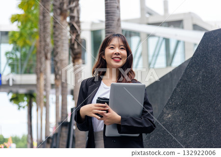 Young Asian business woman, busy professional, wears suit, stands on street, carrying laptop, coffee for work, exudes confidence, smiles brightly in modern urban environment. 132922006